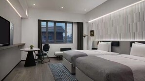Quarto de hotel moderno com duas camas, uma mesinha, uma cadeira e uma janela grande.