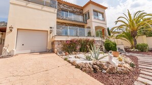 A two-story house with a garage, a balcony, and a landscaped garden with rocks and plants. - Apartment in Roses with Private Pool & Garden (Roses)