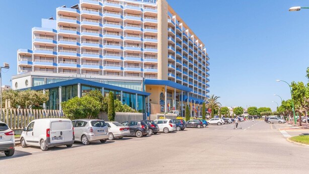 Un hotel de varios pisos con diseño moderno, que cuenta con balcones en cada planta, una estructura vertical central y un estacionamiento con diversos vehículos.