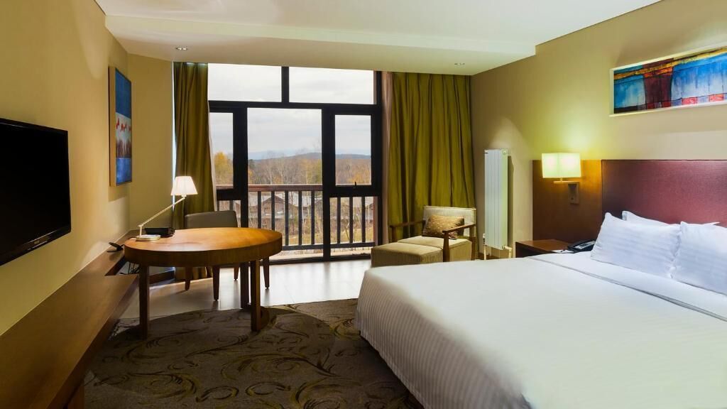 Une chambre d’hôtel avec un lit, un bureau, une chaise, une télévision et une fenêtre avec des rideaux.