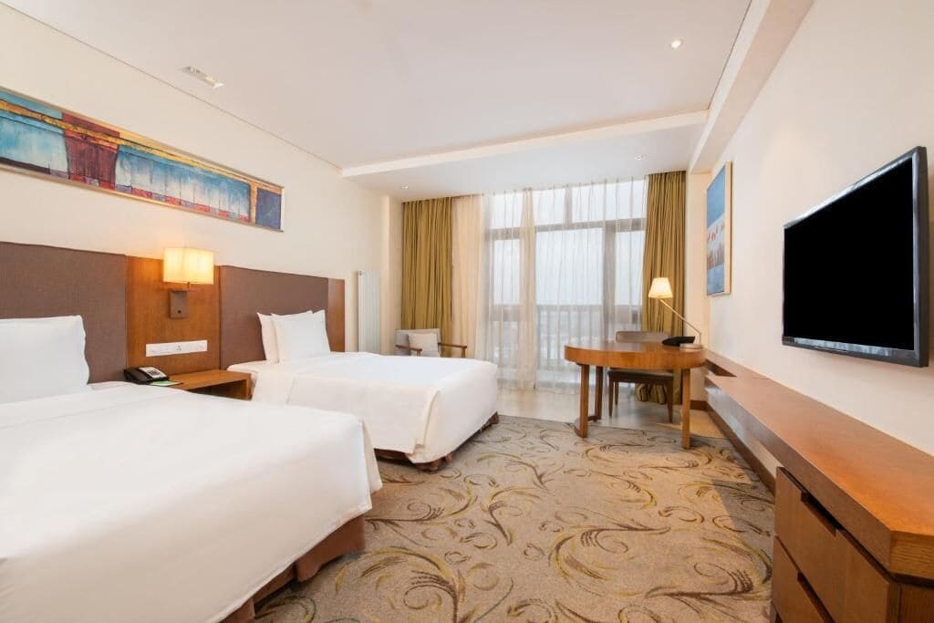 A hotel room with two beds, a desk, a TV, and a large window with curtains.
