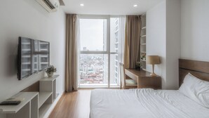 A modern hotel room with a large bed, a TV mounted on the wall, a desk with a lamp, and a view of the city through the window.
