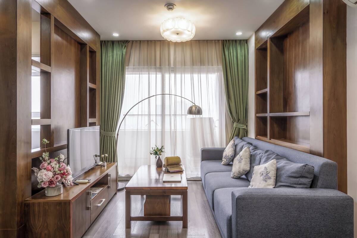 A modern living room with a grey sofa, wooden shelves, a TV, and a coffee table with a vase of flowers.