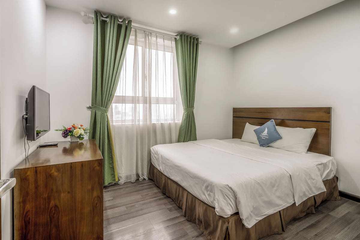 A hotel room with a wooden desk, a bed with white bedding, a blue pillow, and green curtains.