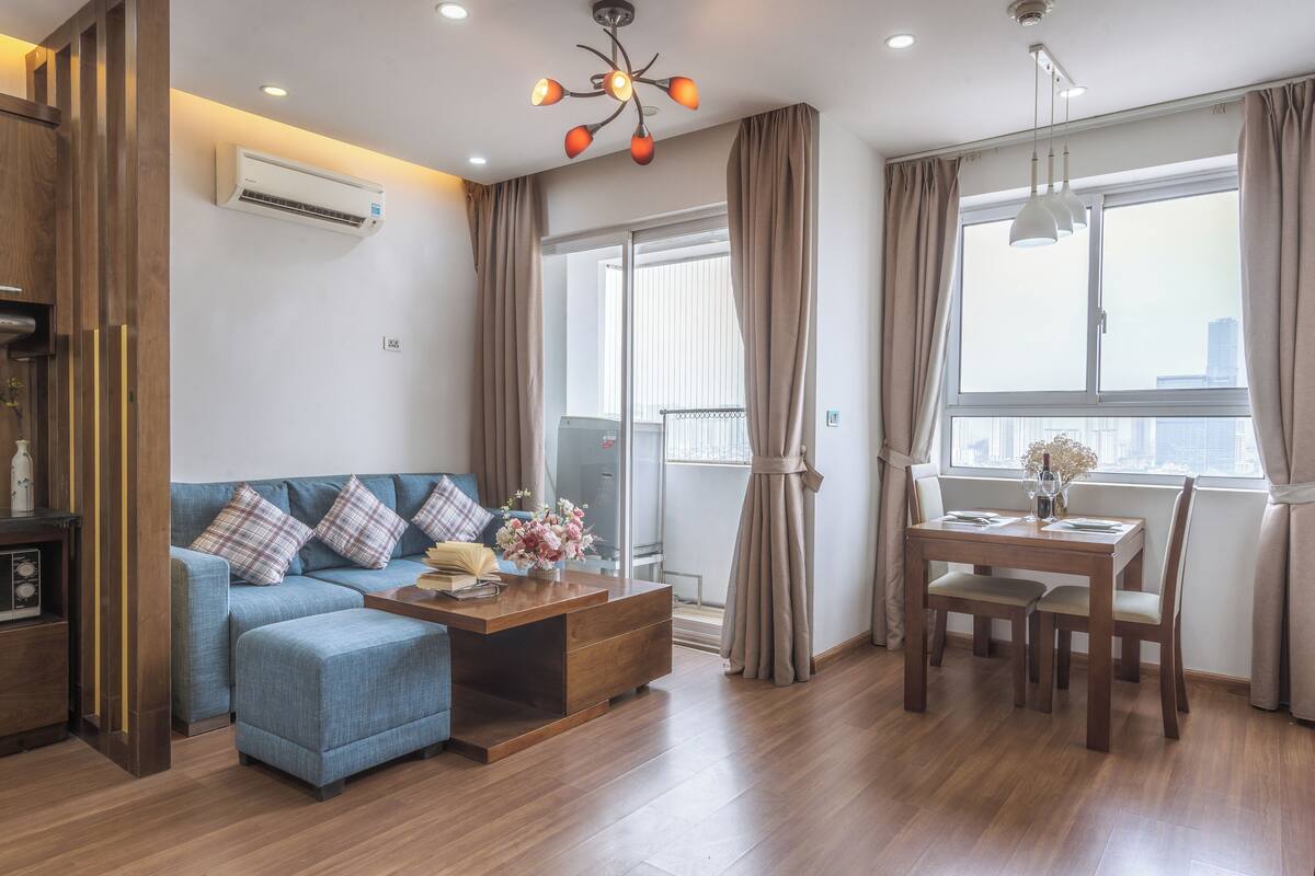 A modern living room with a sofa, ottoman, wooden coffee table, and a dining table with chairs. There is a large window with curtains, an air conditioning unit on the wall, and a ceiling light fixture.
