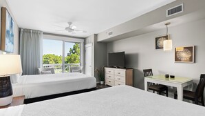 A hotel room with two beds, a desk, a television, and a balcony with a view of trees.