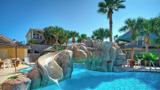 A pool with a water slide, palm trees, and a wooden deck with chairs.