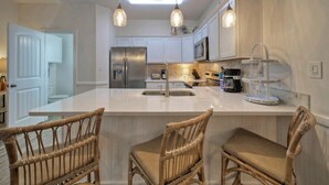 A modern kitchen with white cabinetry, a stainless steel refrigerator, a microwave, and a coffee maker. There are two wicker chairs at the island counter.