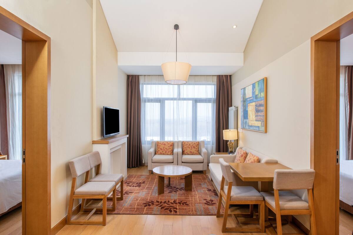 Une chambre d’hôtel moderne équipée d’un canapé, de fauteuils, d’une table à manger, d’un téléviseur et d’une grande fenêtre avec des rideaux.