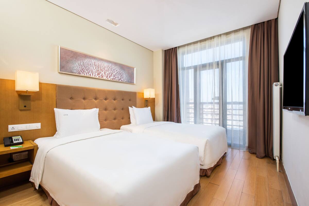 Une chambre d’hôtel avec deux lits, une grande fenêtre avec des rideaux, un téléviseur à écran plat et une œuvre d’art accrochée au mur.