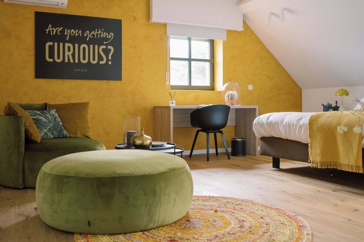 Un cuarto acogedor con un sofá verde, un otomanillo redondo, un escritorio con una silla, una cama con un edredón amarillo y una ventana con vistas.