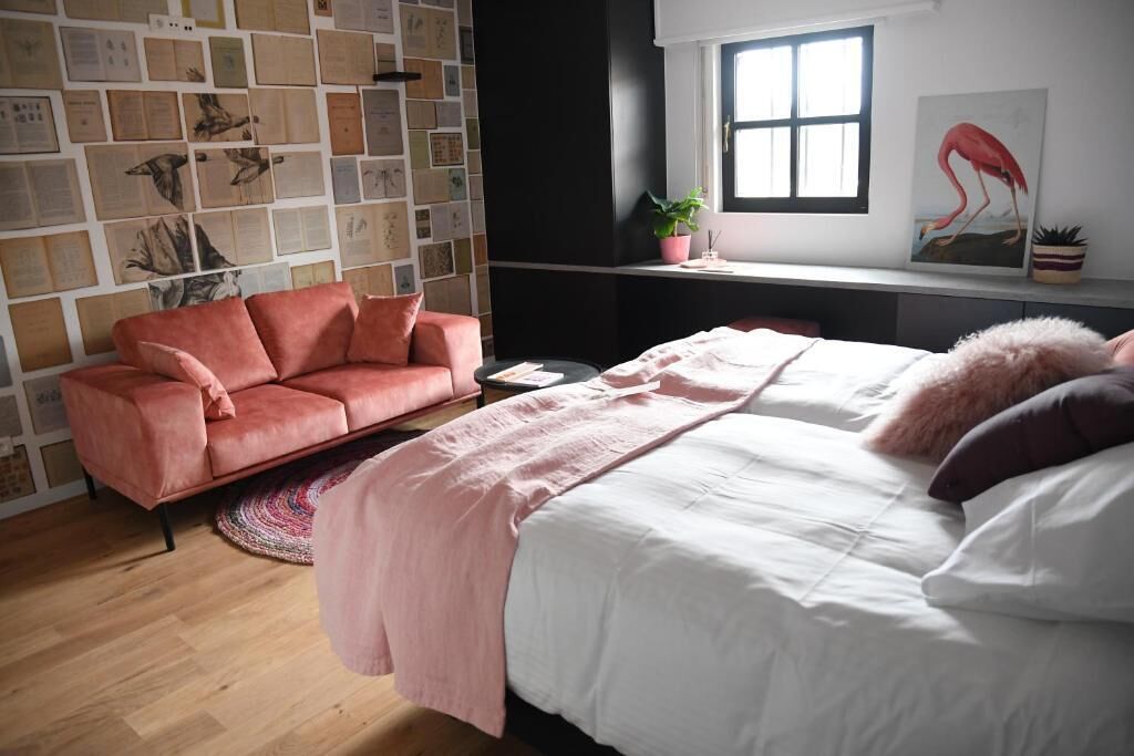 A bedroom with a bed, a pink sofa, a round rug, a window with a view, and a wall decorated with papers.