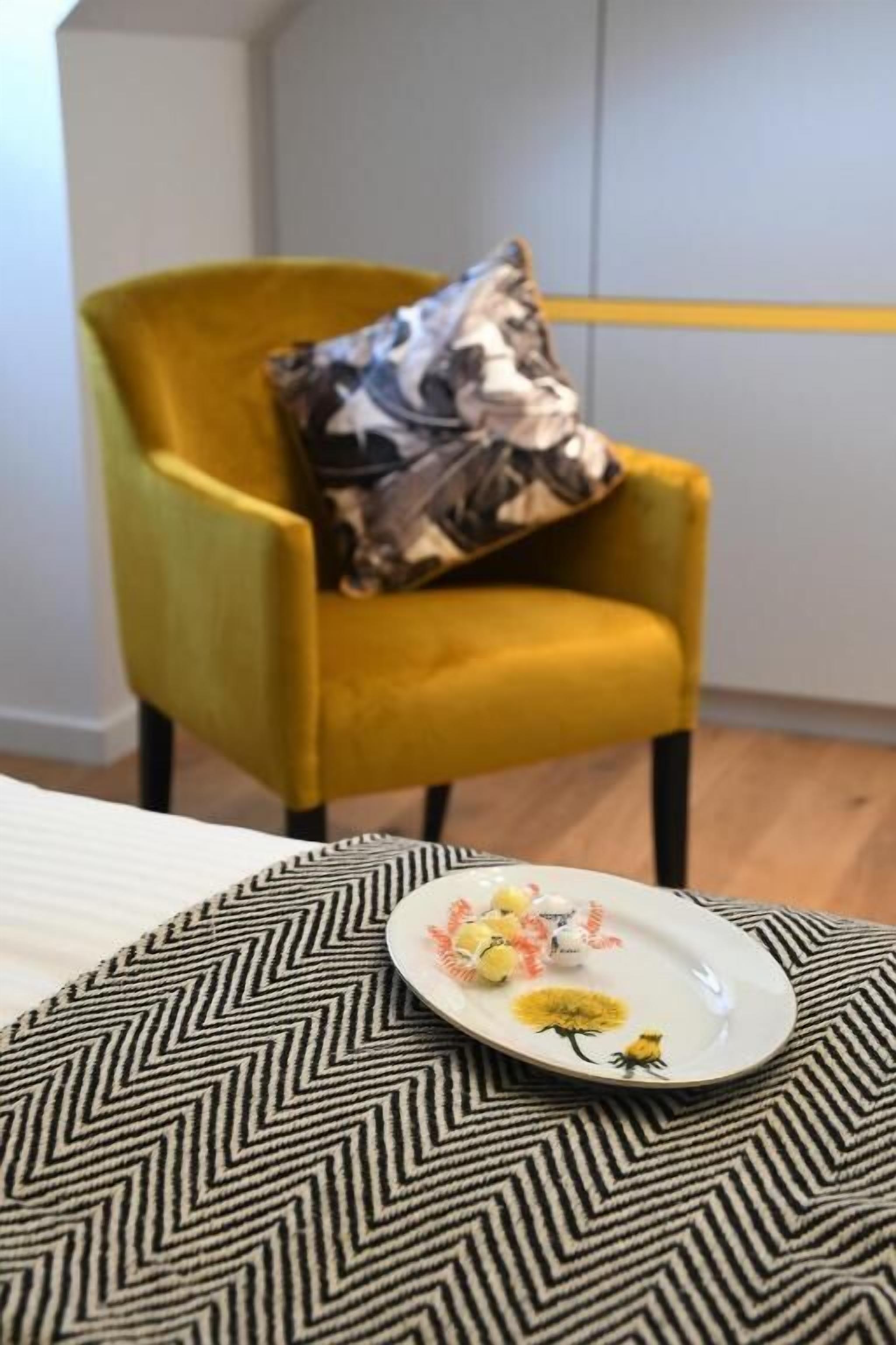 A yellow armchair with a patterned pillow, a plate with decorative items, and a chevron-patterned pillow on a bed.