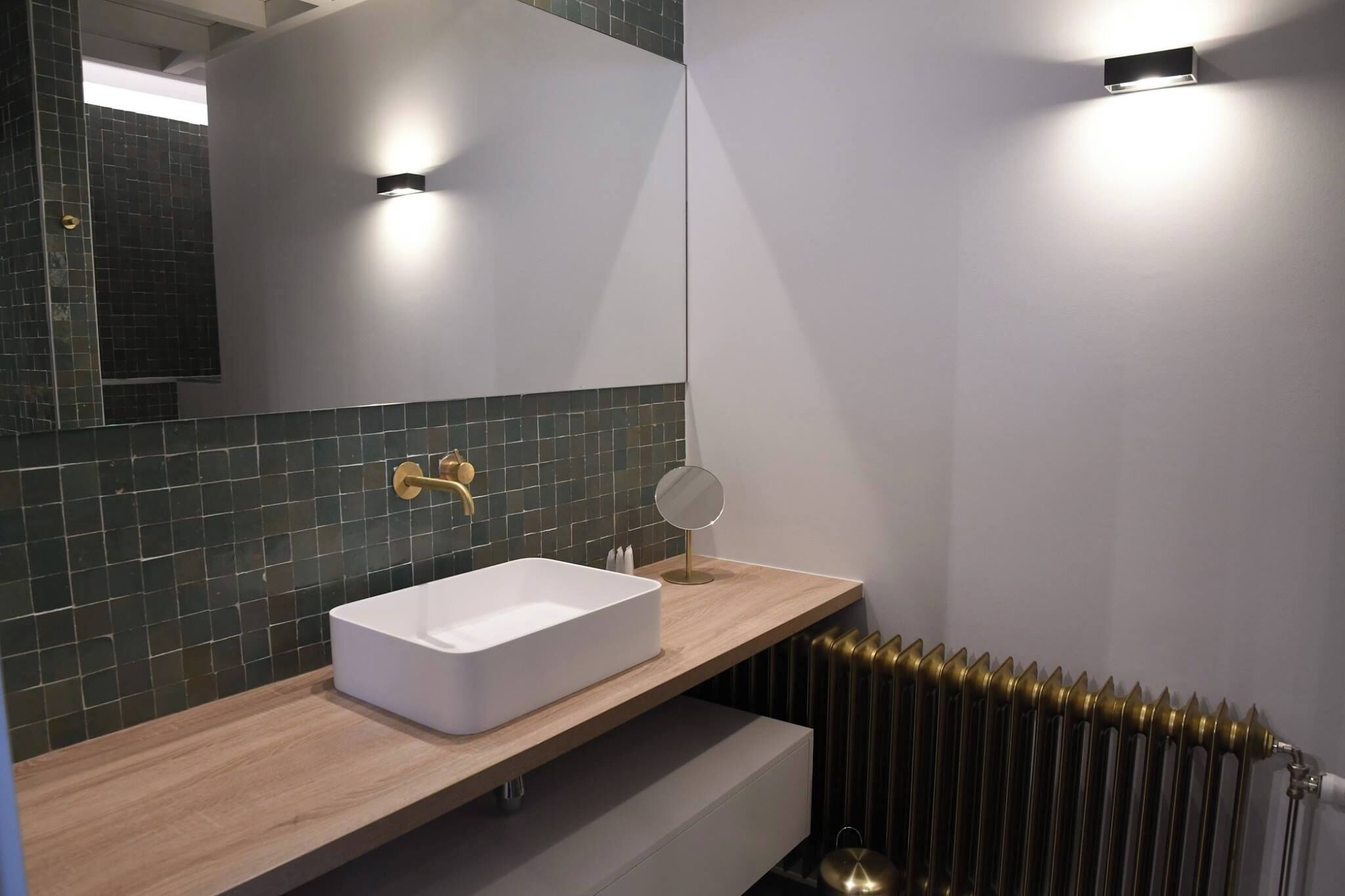 A modern bathroom with a rectangular white sink, a gold faucet, a round mirror, and a radiator.