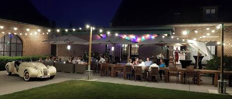 An outdoor patio area with brick walls, string lights, and umbrellas providing shade. There is a vintage car parked in the foreground.