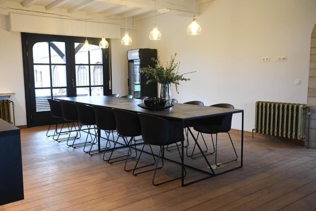 A modern dining area with a long table, black chairs, pendant lights, and a vase with greenery.