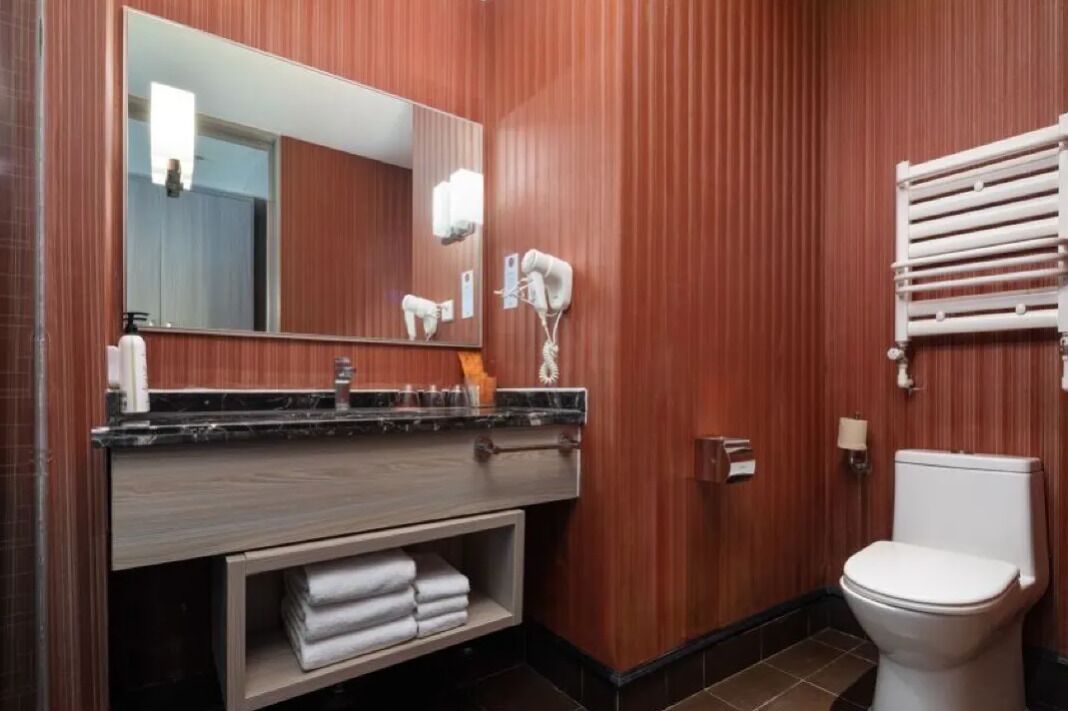 A modern bathroom with a wooden panel wall, a large mirror, a white toilet, a towel rack, and a vanity with a marble countertop.
