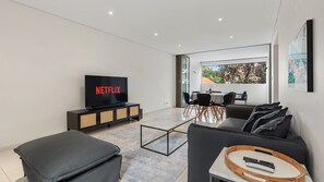 A modern living room with a flat-screen TV, a sofa, a coffee table, and a dining area with chairs and a table.
