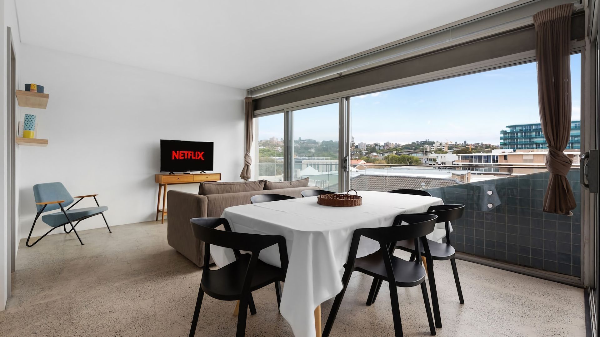 A modern living room with a flat-screen TV displaying 'NETFLIX,' a dining area with a table and chairs, and a large window offering a city view.