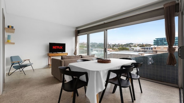 A modern living room with a flat-screen TV displaying 'NETFLIX,' a dining area with a table and chairs, and a large window offering a city view.