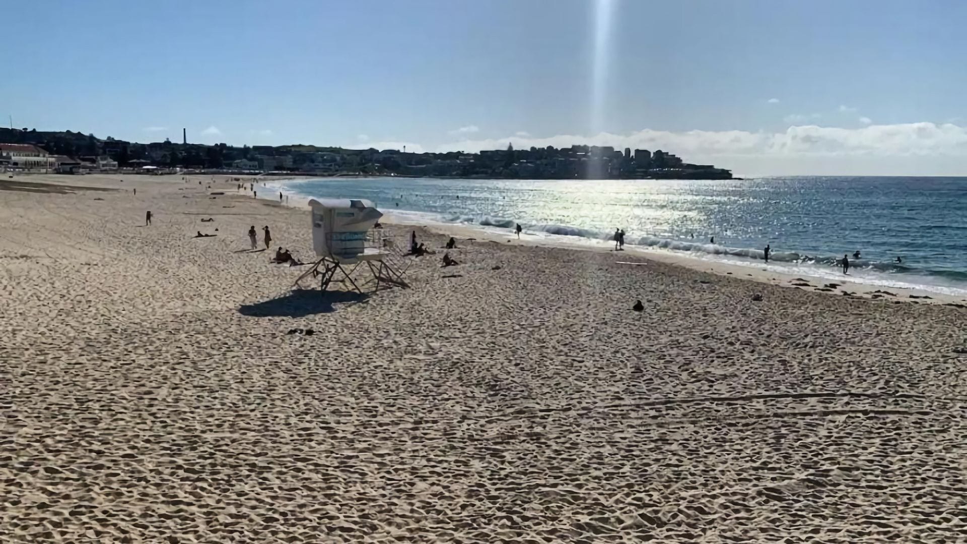 Una playa arenosa con una torre de salvavidas, olas del océano y una costa distante con edificios.