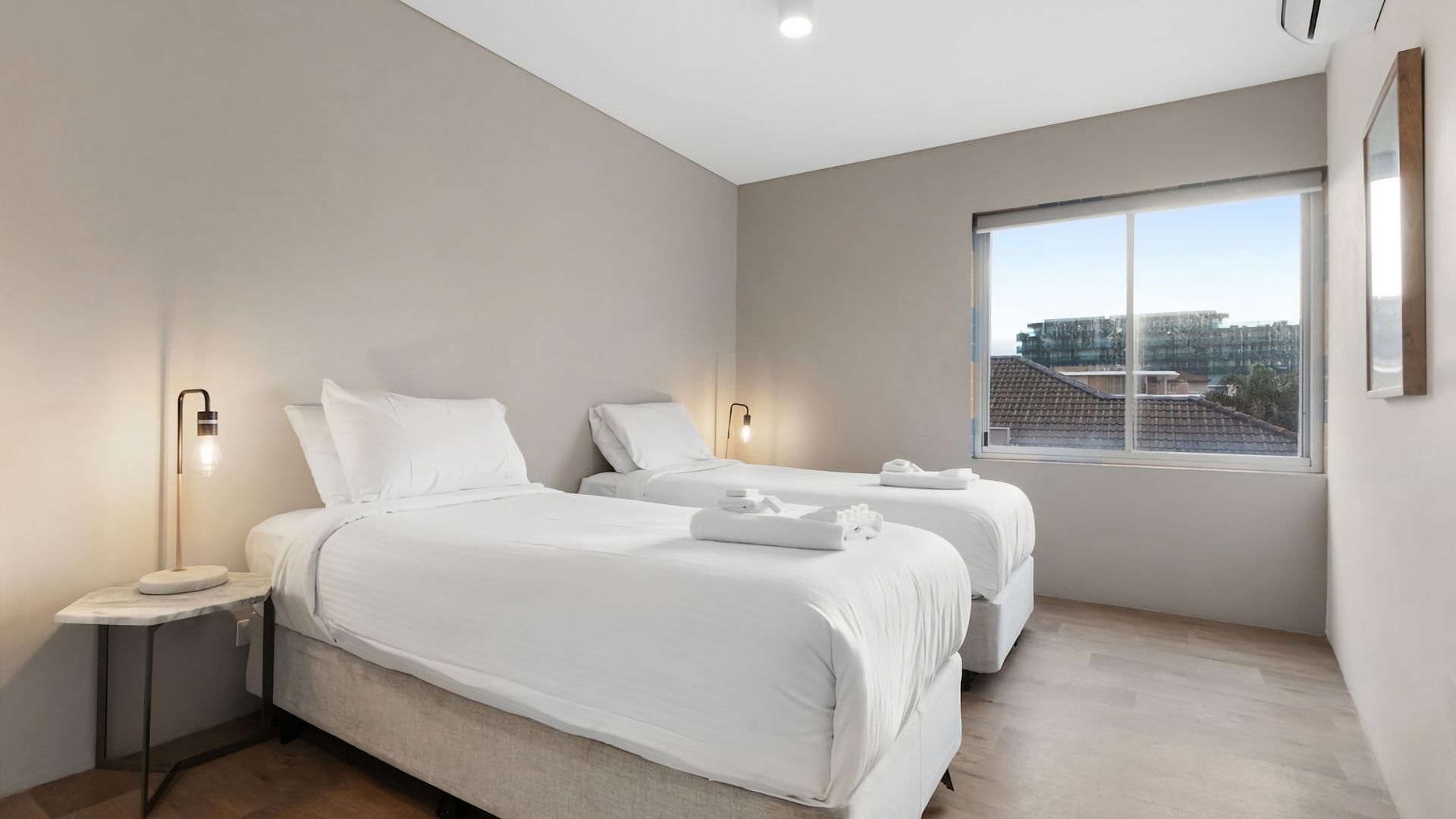 A modern hotel room with two beds, a nightstand, a bedside lamp, and a large window offering a city view.