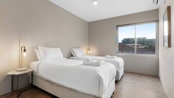 A modern hotel room with two beds, a nightstand, a bedside lamp, and a large window offering a city view.
