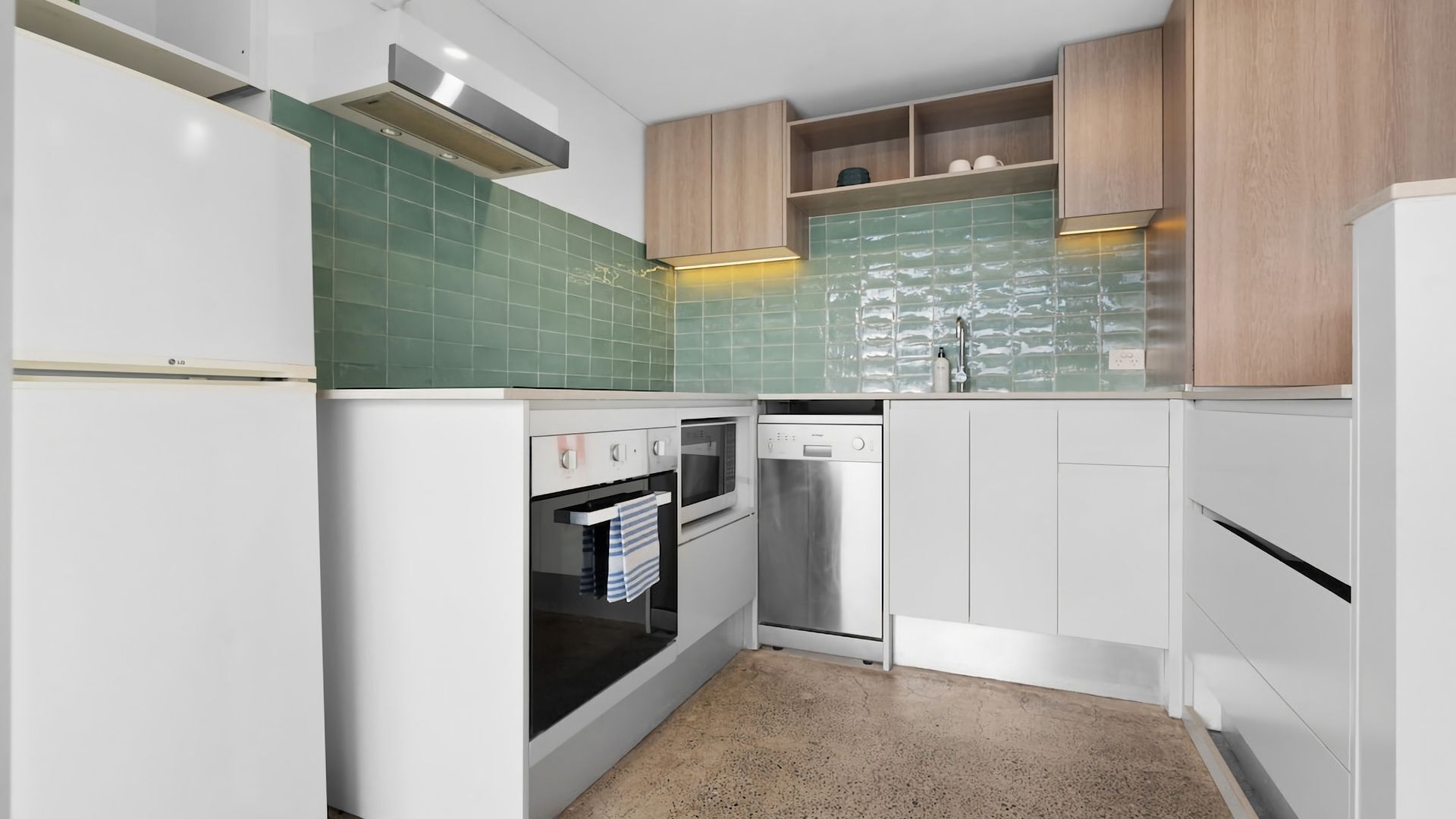 Una cocina moderna con armarios blancos, salpicadero de azulejos verdes y electrodomésticos de acero inoxidable, incluyendo lavavajillas y horno.