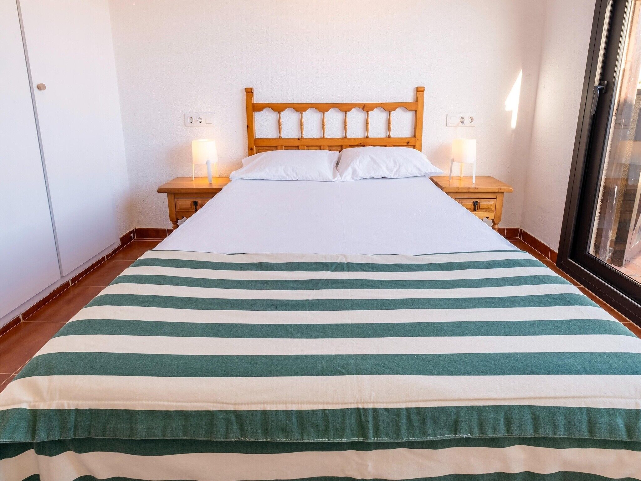 Un letto con copriletto a righe, testiera in legno, due comodini con lampade e una finestra con persiane.