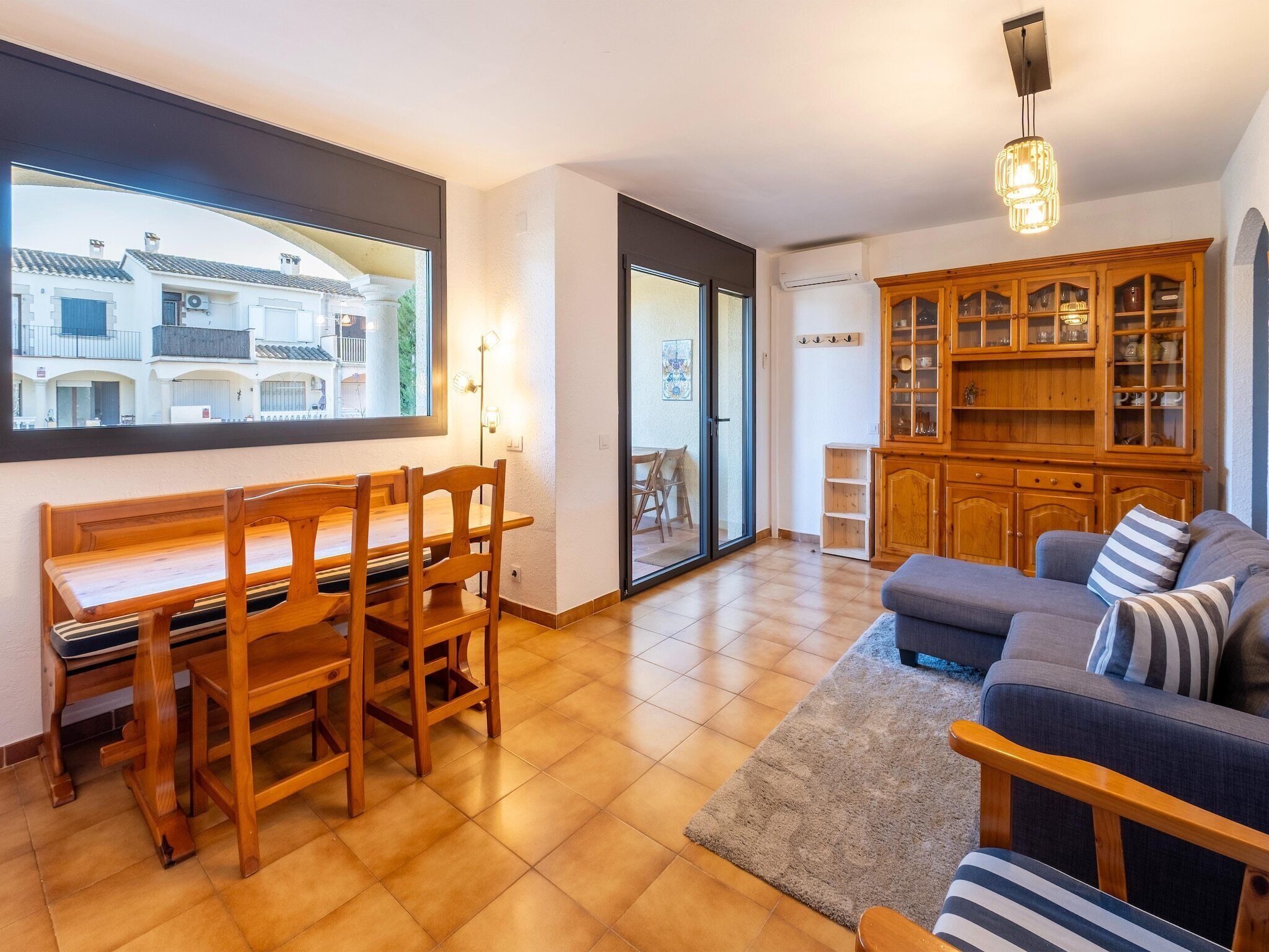 A spacious living room with a wooden dining table and chairs, a grey sofa, and a wooden cabinet with glass doors.