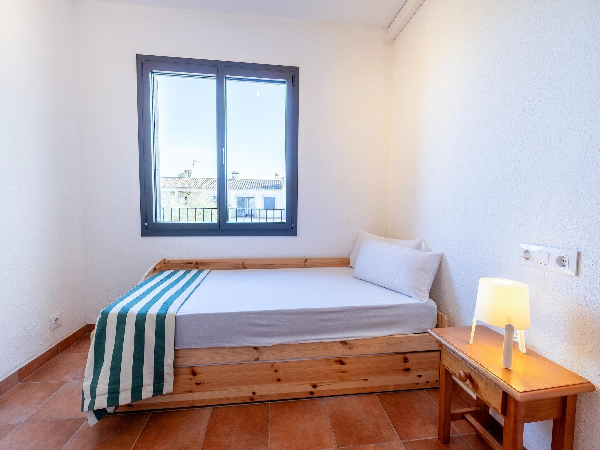 Een kleine kamer met een enkel bed, houten bedframe, een raam met uitzicht op gebouwen, een nachtkastje met een lamp en een stopcontact aan de muur.