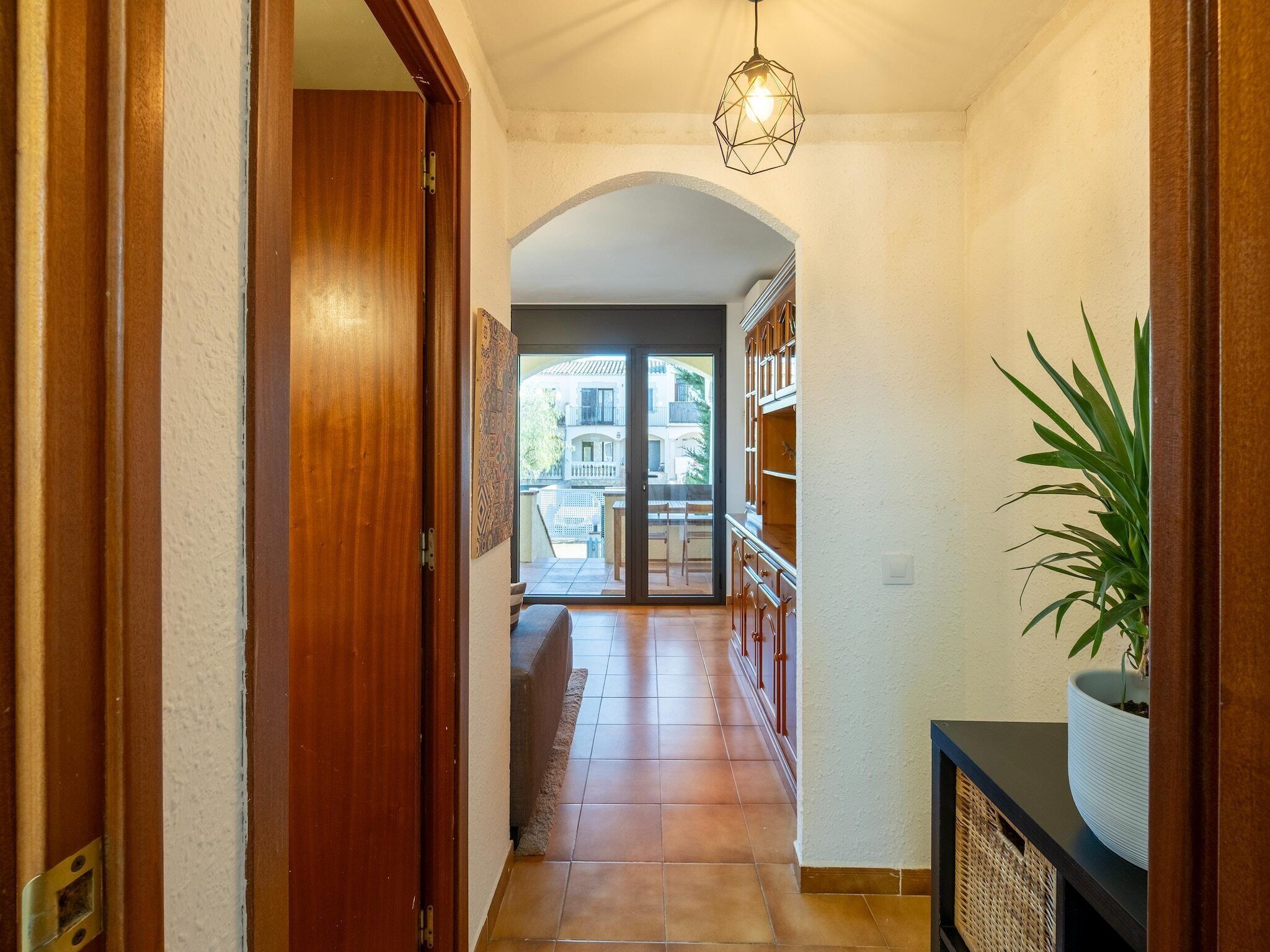 Un corridoio con una porta in legno, pavimento piastrellato, lampadario geometrico e una pianta in vaso.