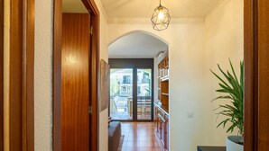 Un pasillo con puerta de madera, suelo de baldosas, lámpara con diseño geométrico y una planta en maceta.
