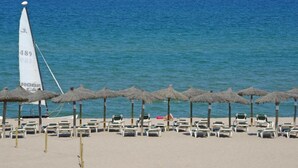 Ein Strand mit Liegestühlen und reetgedeckten Sonnenschirmen, ein Segelboot in der Ferne und ein klares, blaues Meer.