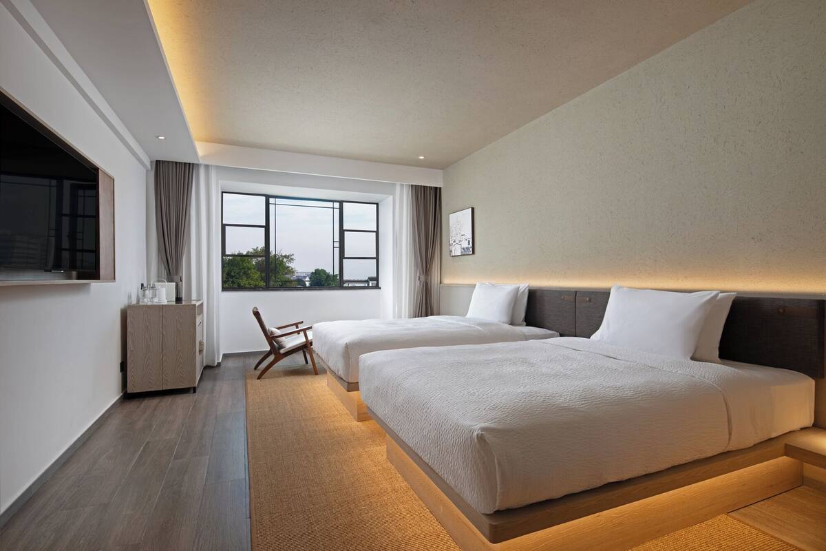 A modern hotel room with two beds, a large window, a flat-screen TV, and a small table.