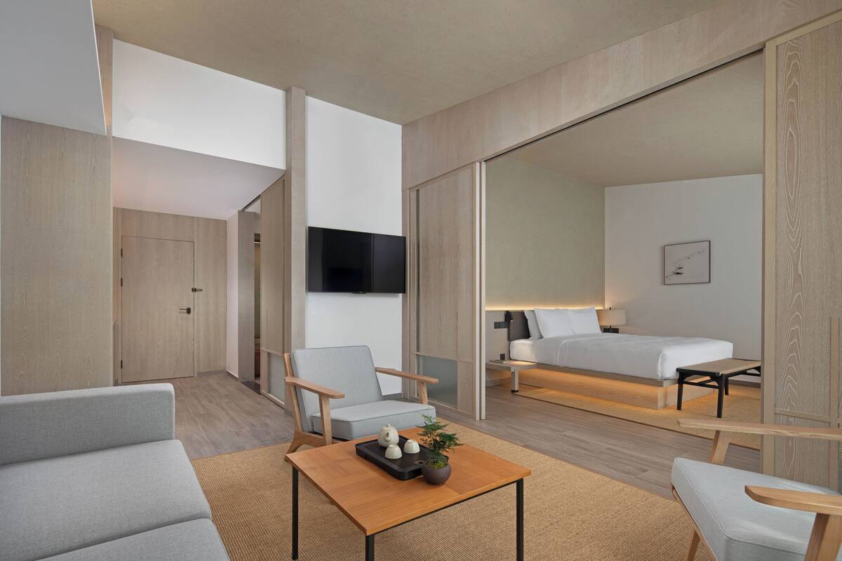 A modern hotel room with a sofa, armchair, and a small table with decorative items. There is a bed with a bedside table and a lamp, and a TV mounted on the wall.
