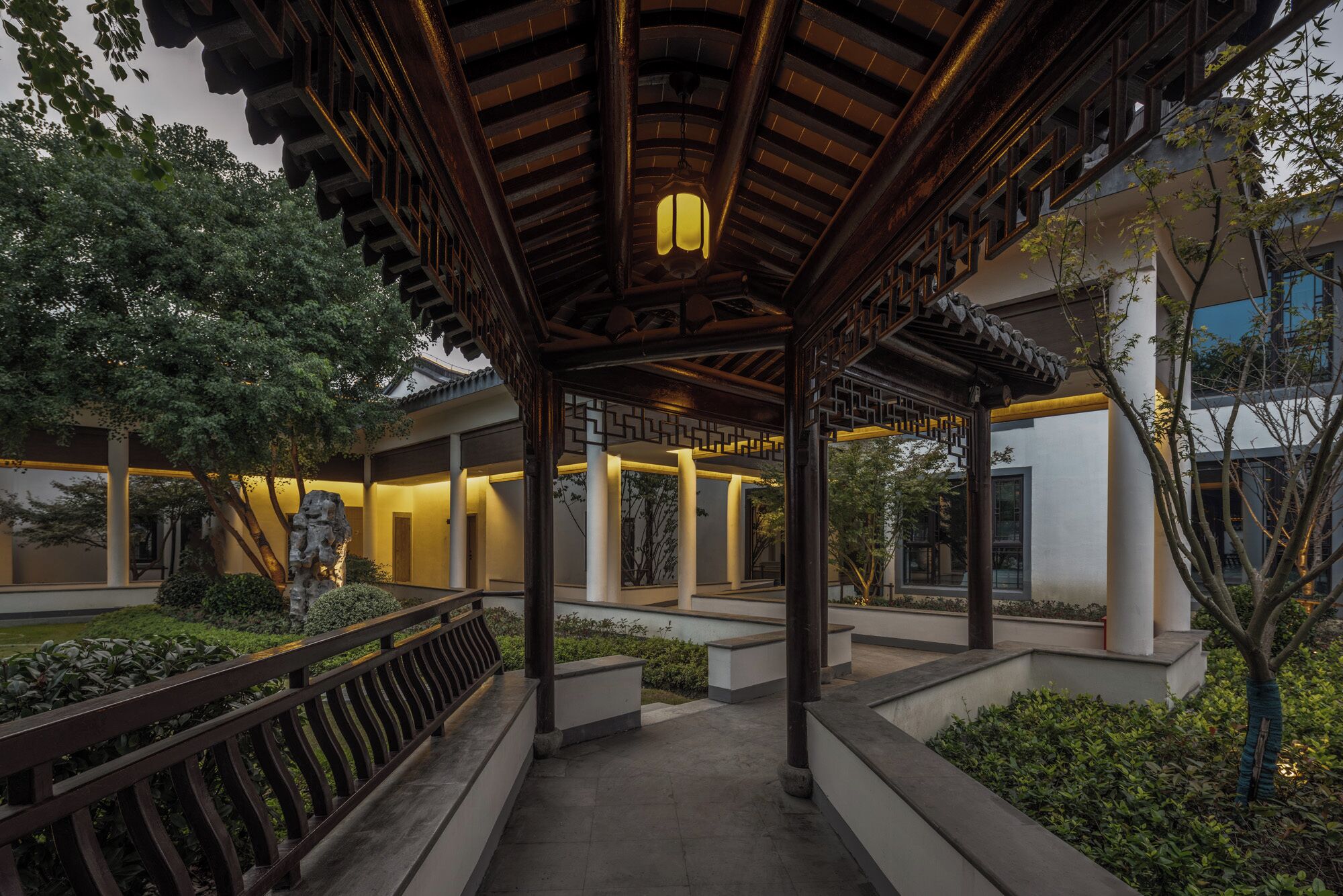 A traditional-style pavilion with a wooden roof and lanterns, surrounded by greenery and modern buildings.