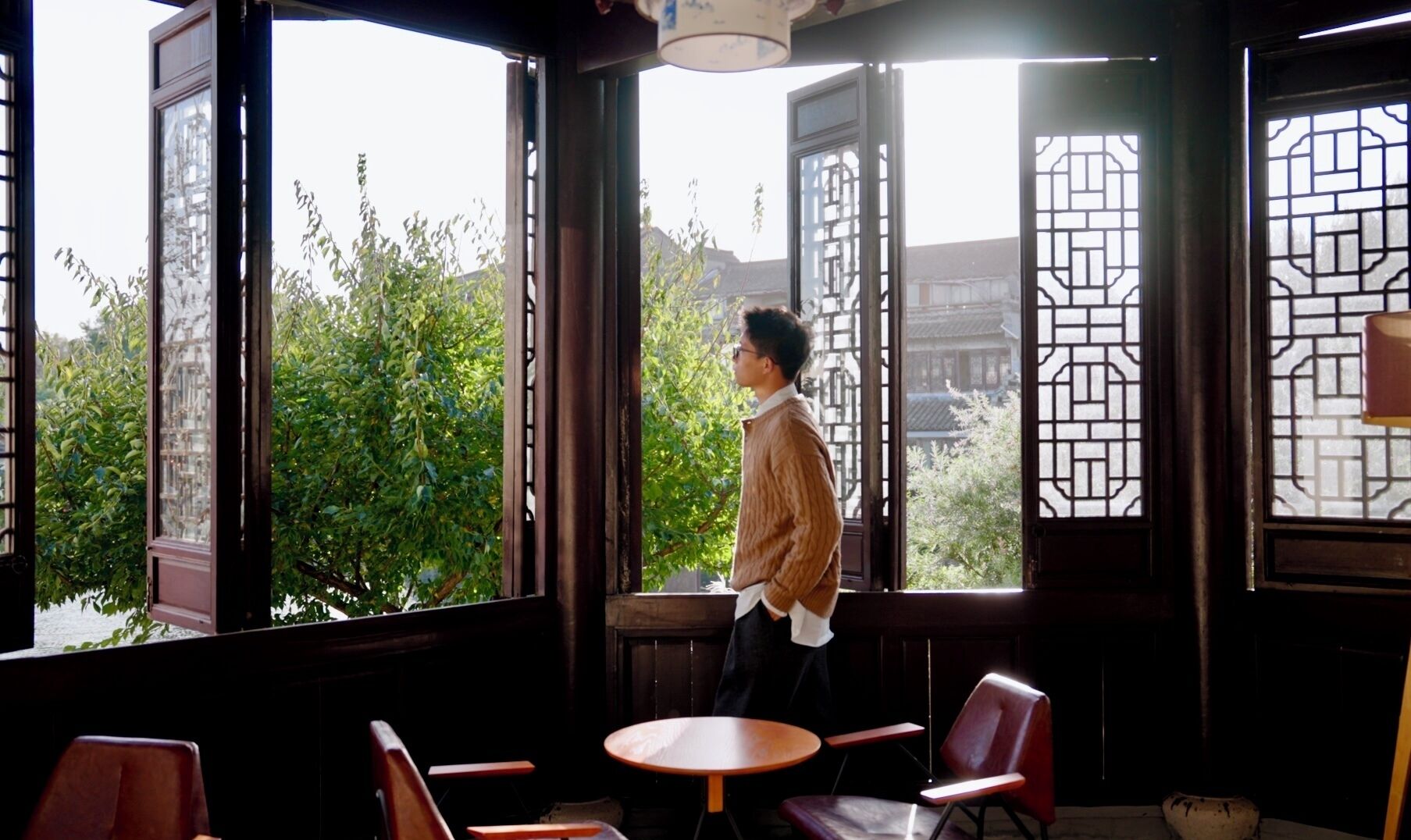 A person standing by a window with traditional lattice design, overlooking greenery and a building. Inside, there are wooden chairs and a small round table.