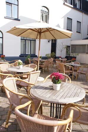Relax in the serene outdoor seating area with a comfortable table and wicker chairs, surrounded by lush greenery and a large umbrella.