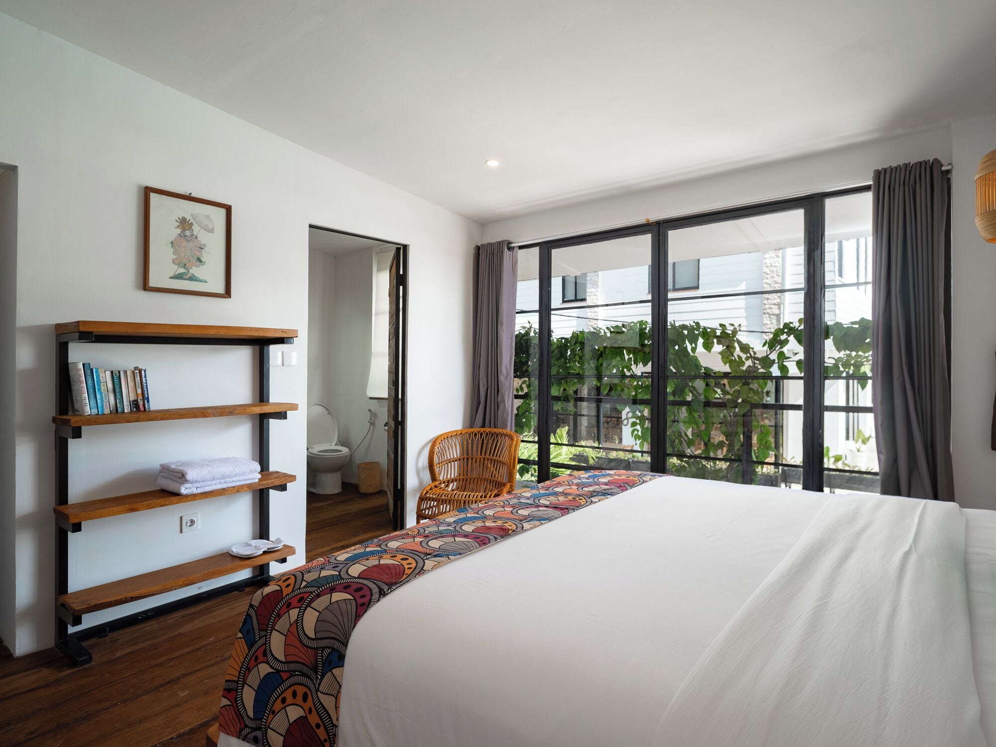 A bedroom with a large bed, a wooden bookshelf, a wicker chair, and a view of greenery through large windows.