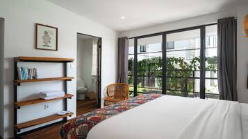 A bedroom with a large bed, a wooden bookshelf, a wicker chair, and a view of greenery through large windows.