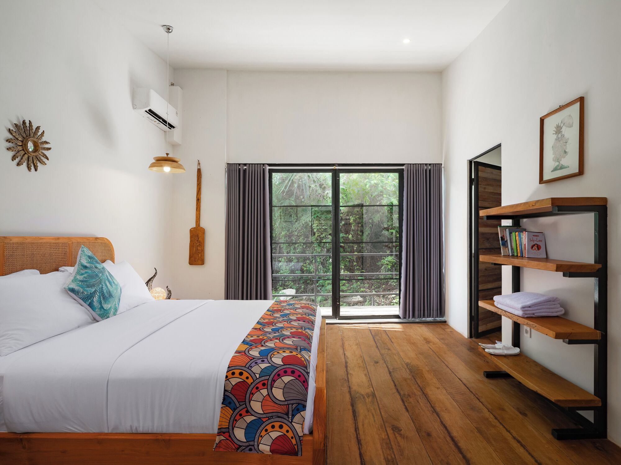 A bedroom with a bed, a wooden bench, a shelf with books, and a view of greenery through the window.