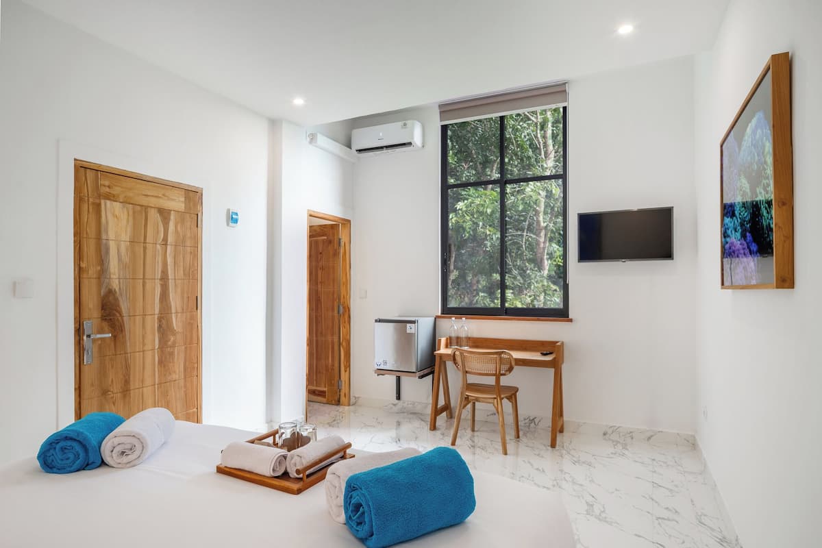 A modern bedroom with a large bed, a desk, and a window with a forest view.