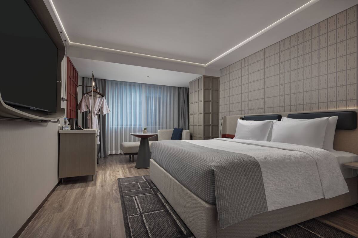 A modern hotel room with a large bed, a TV, a desk, and a seating area.