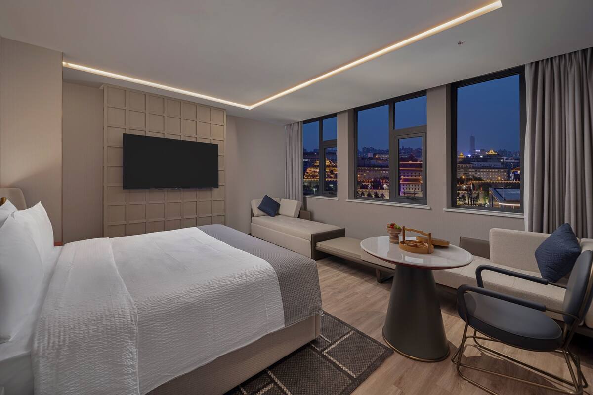 A modern hotel room with a large bed, a flat-screen TV, a sofa, a small round table, and a chair.