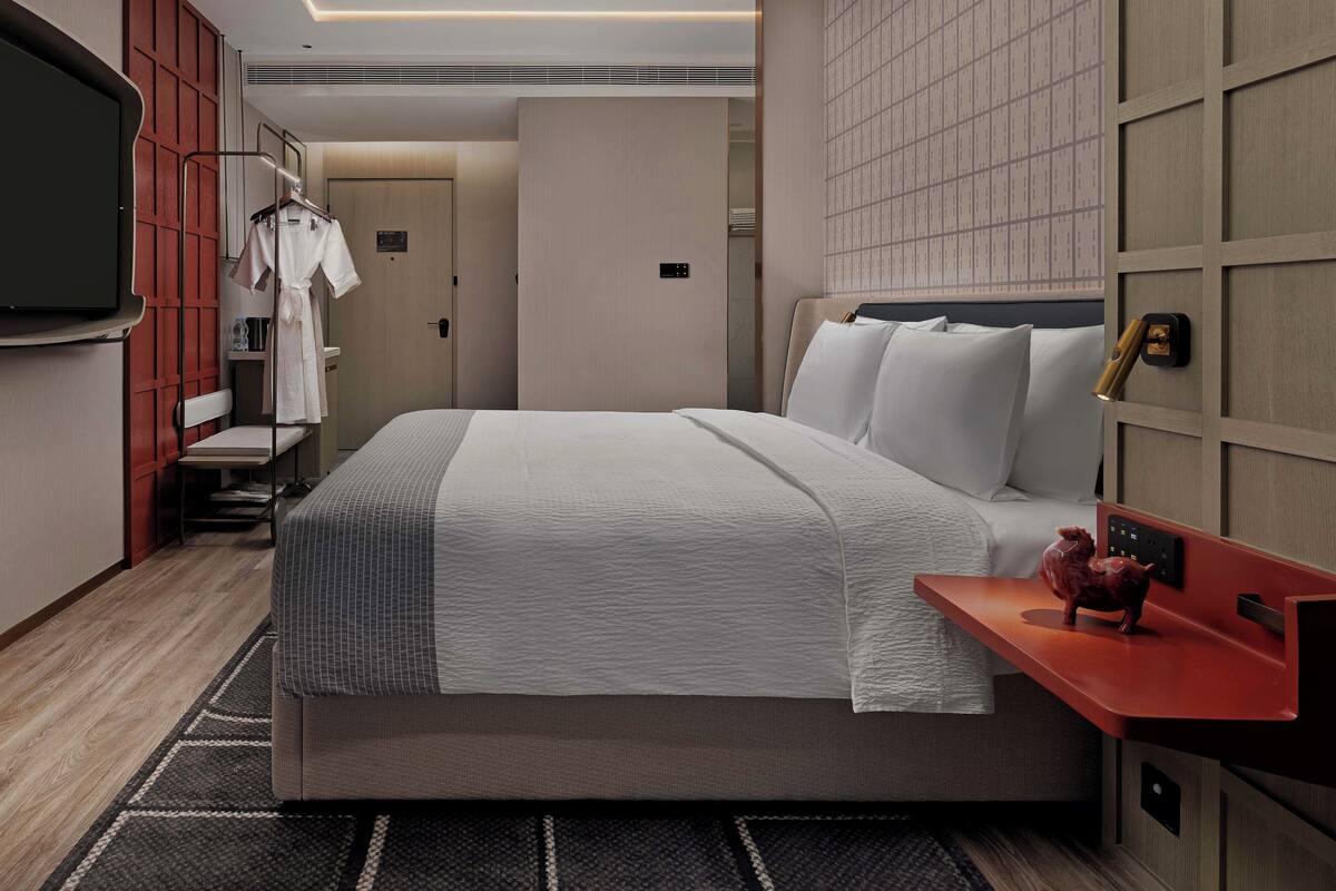 A modern hotel room with a large bed, a flat-screen TV mounted on the wall, a red side table with a decorative item, and a wardrobe with a mirror.