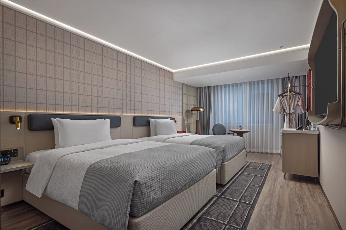 A modern hotel room with two beds, a bedside table with a lamp, a small table with a chair, and a large mirror.