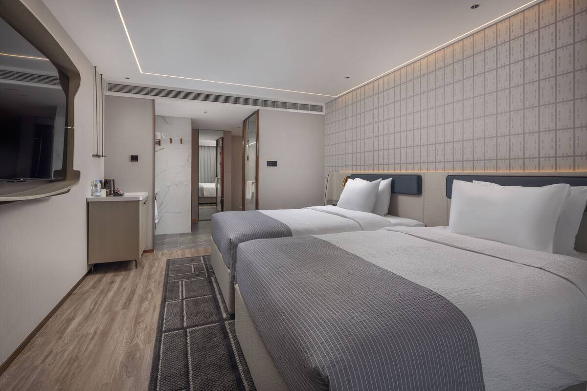 A modern hotel room with two beds, a small desk, a mirror, and a bathroom visible through an open door.