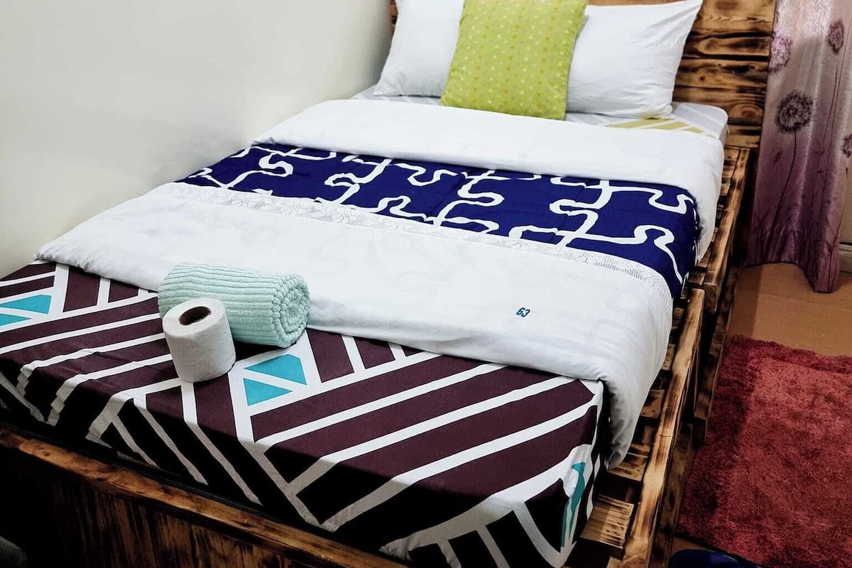 A single bed with a patterned bedspread, pillows, and a rolled-up towel.