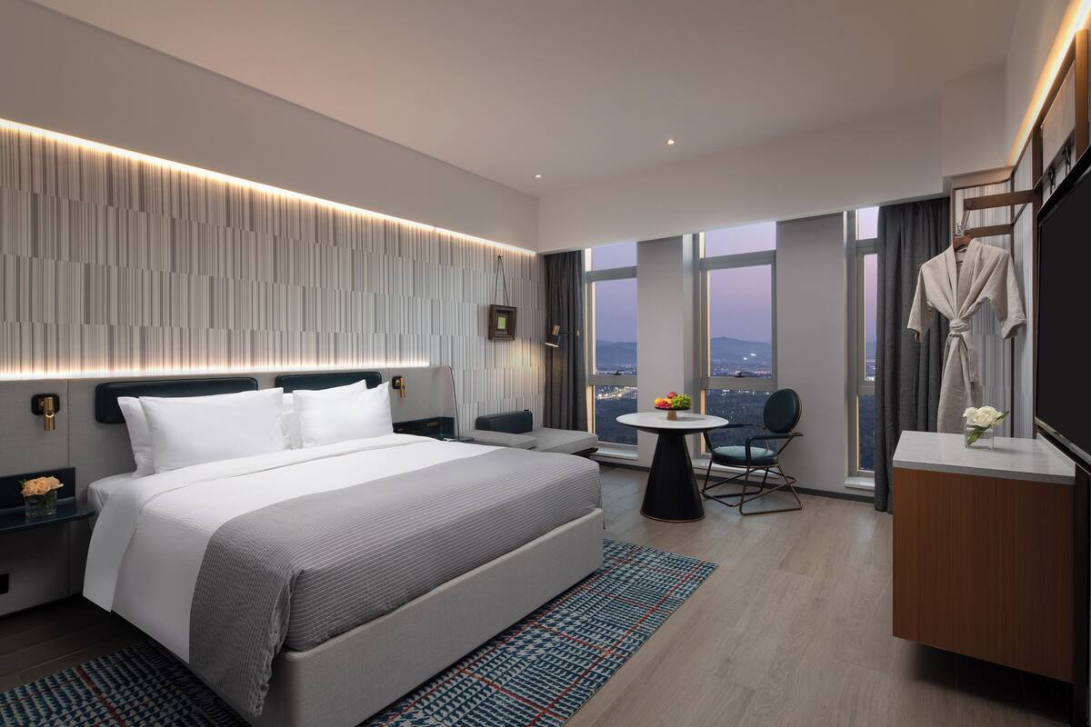 A modern hotel room with a large bed, a small round table, a seating area, and a view of the city.
